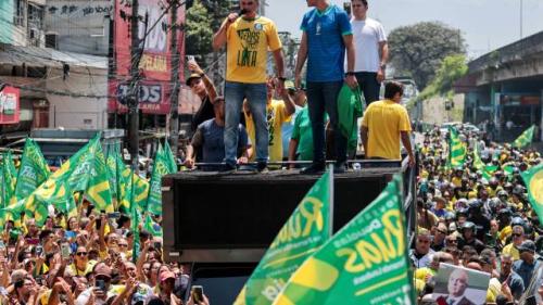Homem de camisa amarela com microfone em cima de um trio elétrico, rodeado por outros homens e bandeiras verdes e amarelas, em meio a uma multidão em manifestação na rua